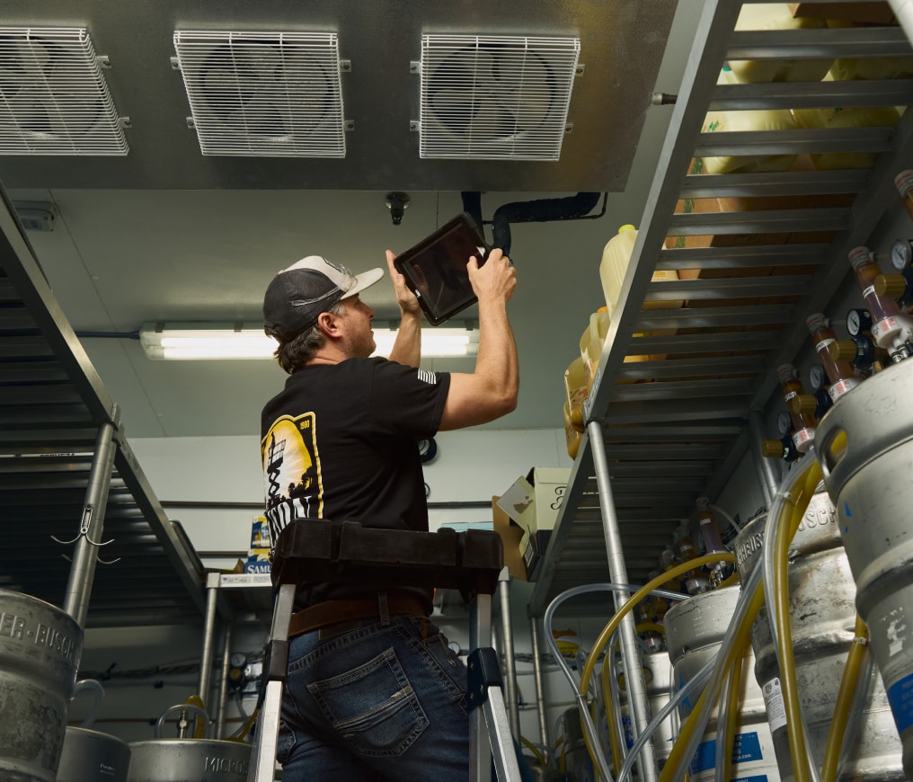 A person in a cap checks equipment inside an industrial room with metal shelves and kegs, using a tablet for reference.