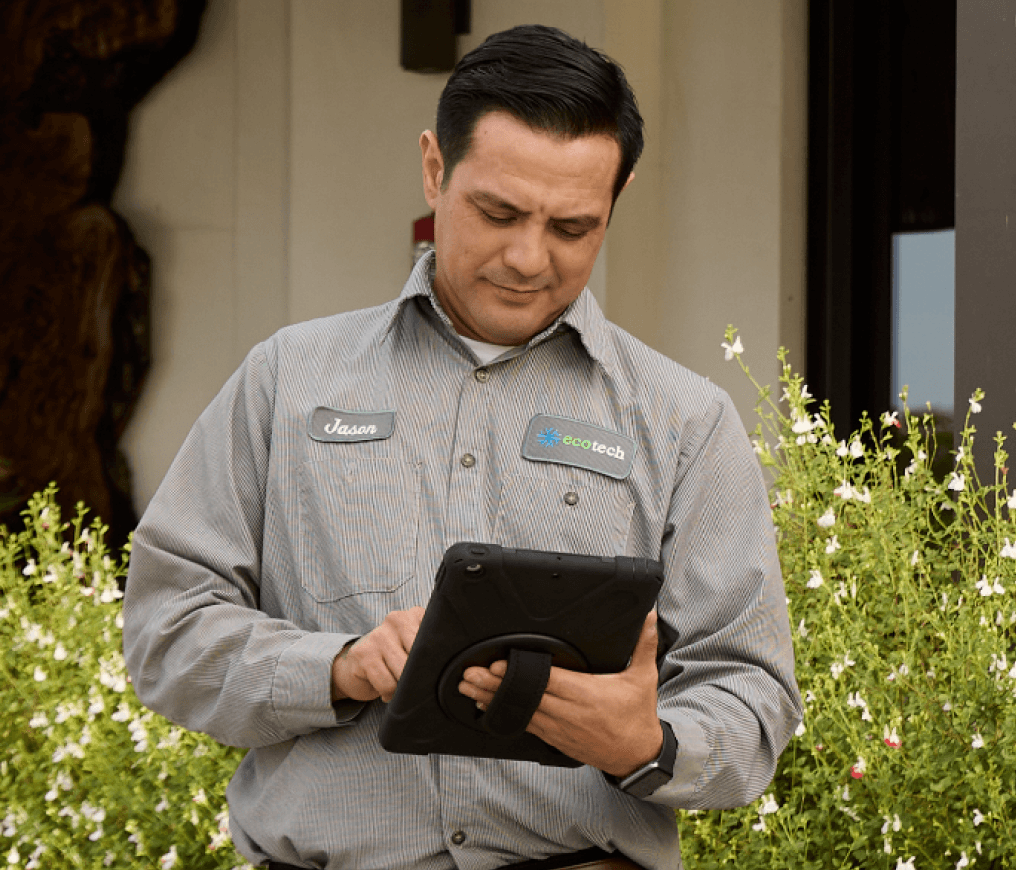 A man in a uniform uses a tablet while standing in front of a building, surrounded by greenery and flowering plants.