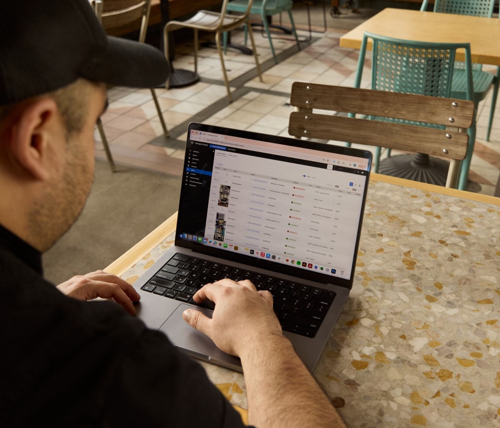 Person in a black shirt and cap using a laptop with spreadsheets on the screen, seated at a speckled table in a cafe.