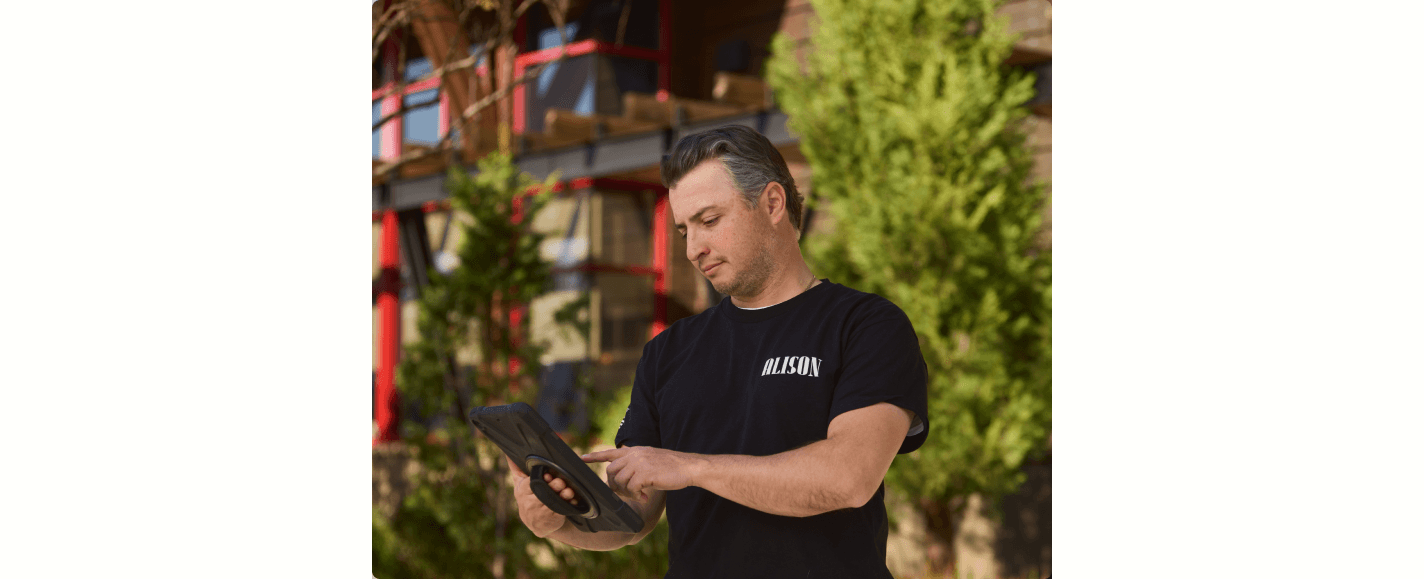 A man in a black "Milason" shirt uses a tablet outdoors, standing in front of a building with greenery around.
