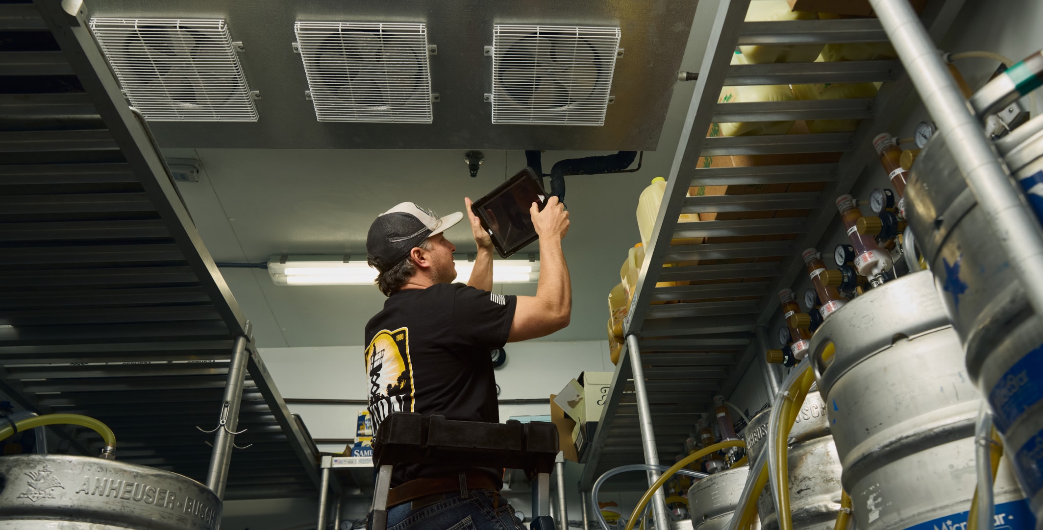 A person in a cap checks equipment inside an industrial room with metal shelves and kegs, using a tablet for reference.