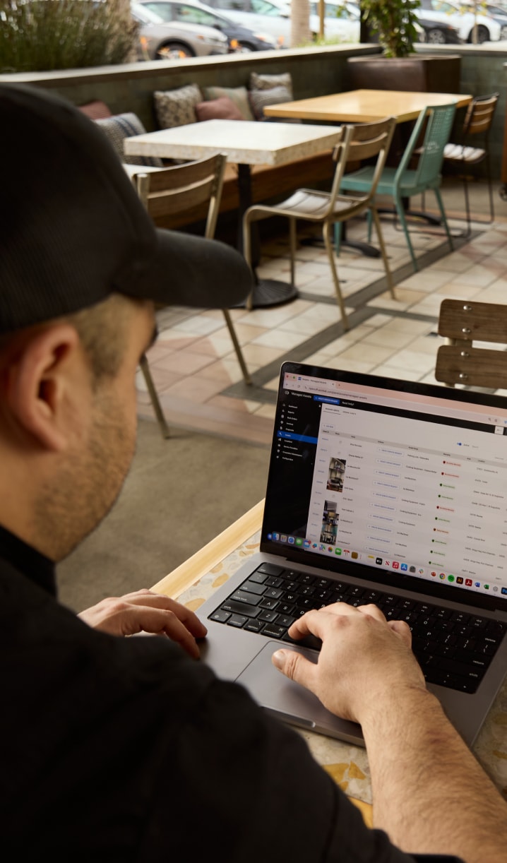 Person sitting at a table, using a laptop in a cafe. The screen displays a list with columns, possibly a spreadsheet or data table.