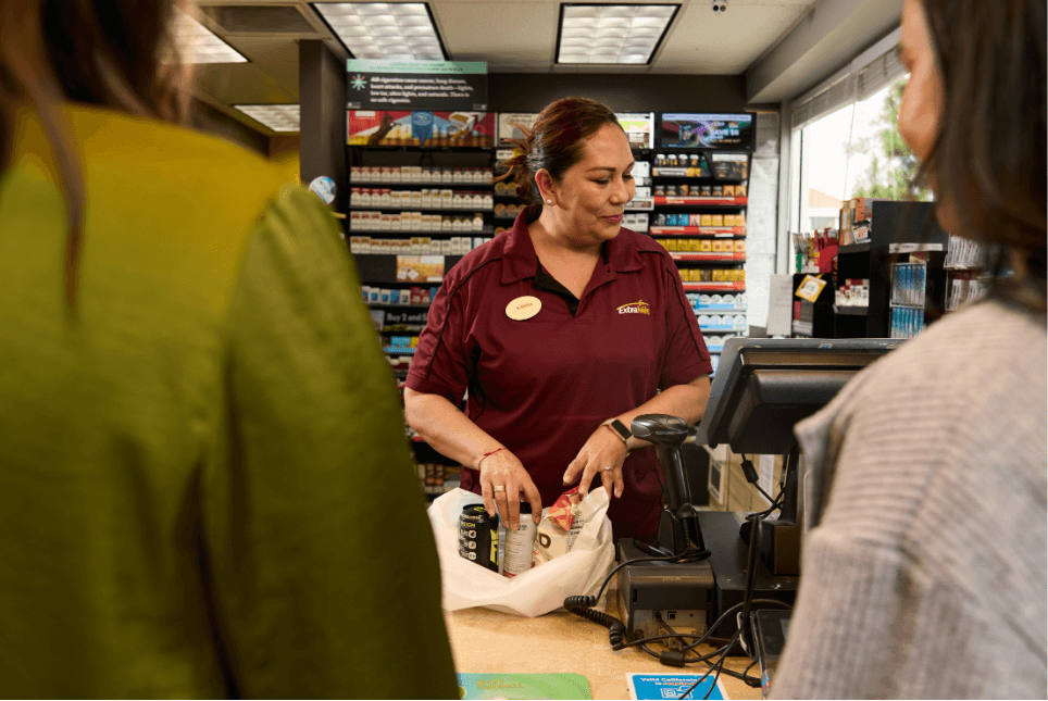 Cashier in a maroon uniform bagging items at a convenience store, with two customers standing at the counter.